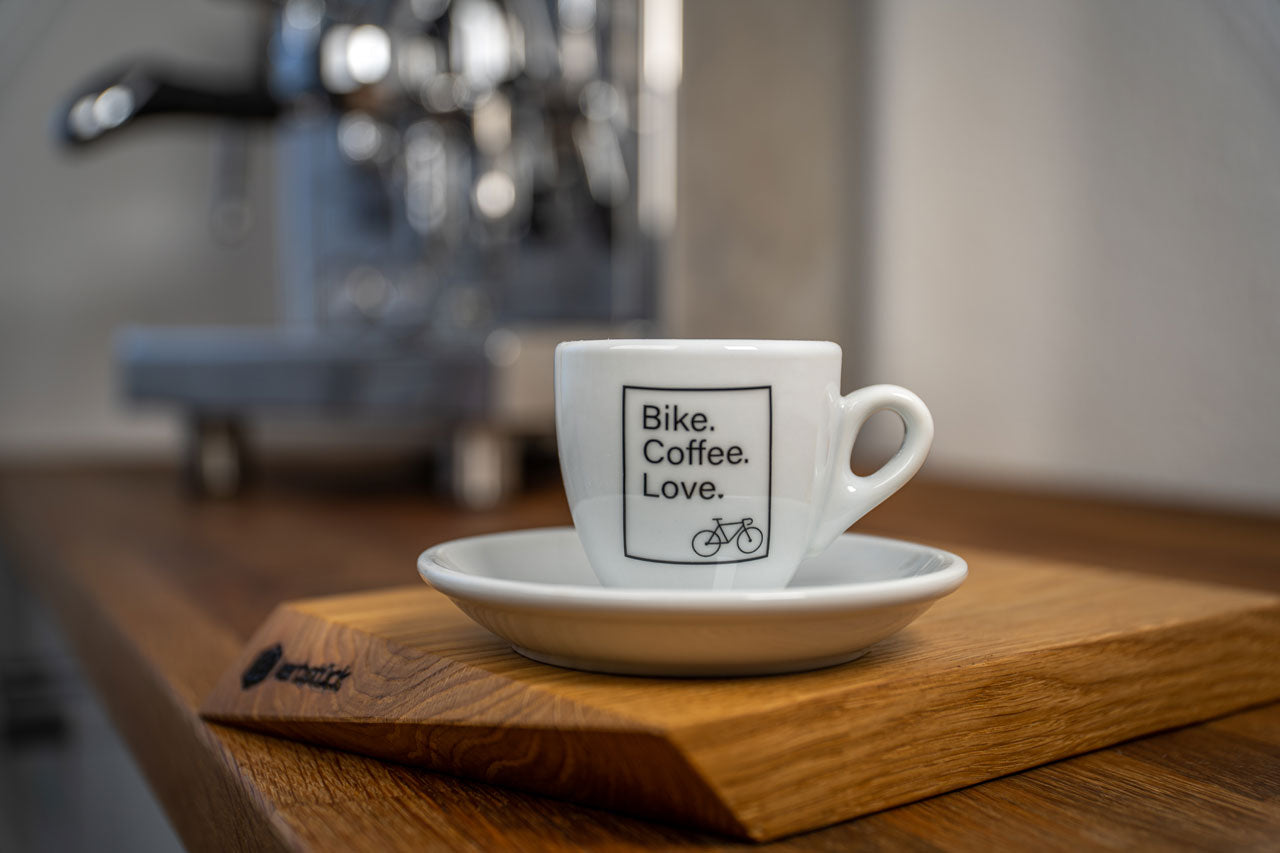 Eine weiße Espresso-Tasse bedruckt mit dem Slogan "Bike Coffee Love" und einem stilisierten Rennrad in einem schwarzen Kasten.