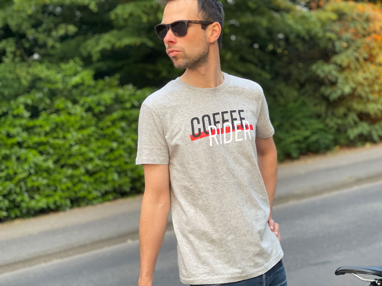 Eine Person steht mit einem grauen T-Shirt und der Aufschrift Coffee Rider vor einer Hecke