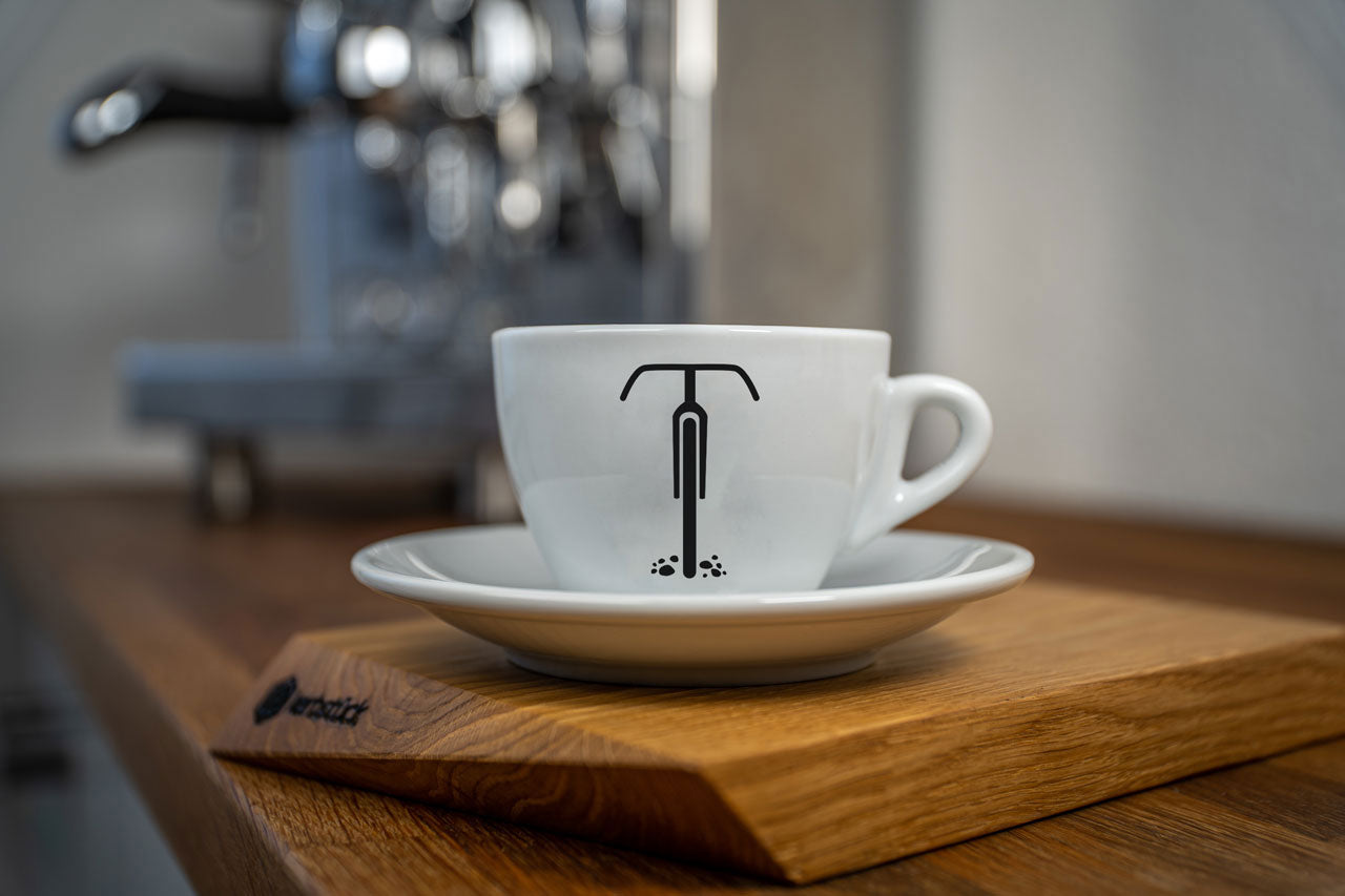Eine weiße Cappuccino-Tasse bedruckt mit der schwarzen Silhouette eines Gravel-Rades. Im Hintergrund eine glänzende Siebträger-Kaffeemaschine.