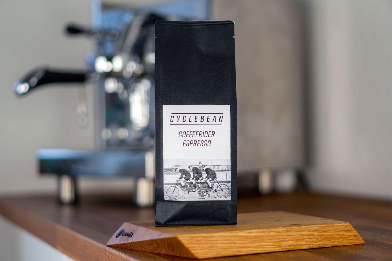 Eine Espresso-Packung steht auf einer Arbeitsfläche vor einer Kaffeemaschine. Auf den Label sind drei Radfahrer zu sehen.