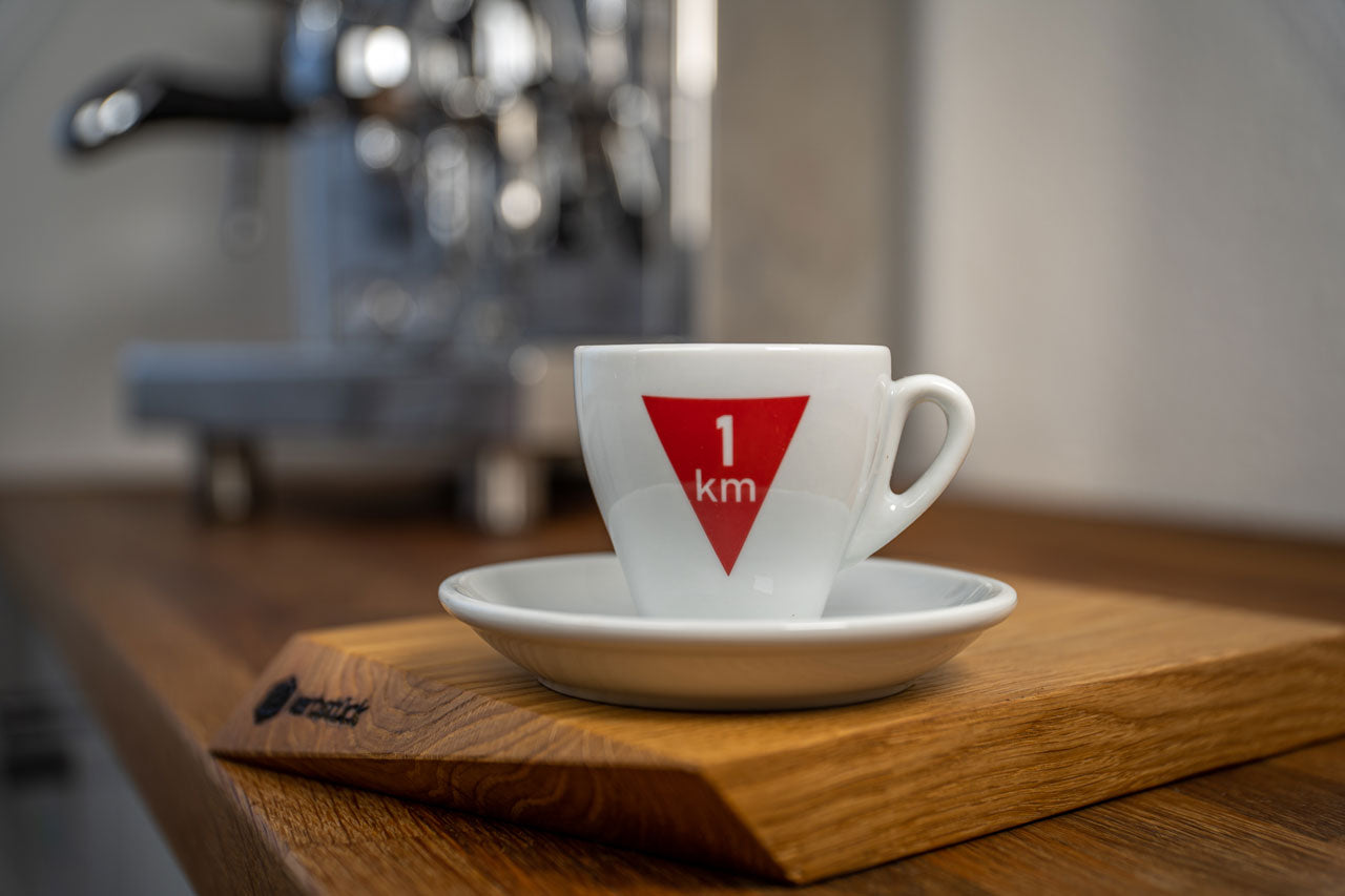 Eine weiße Espresso-Tasse bedruckt mit einem roten umgedrehten Dreieck mit der weißen Aufschrift 1 km.