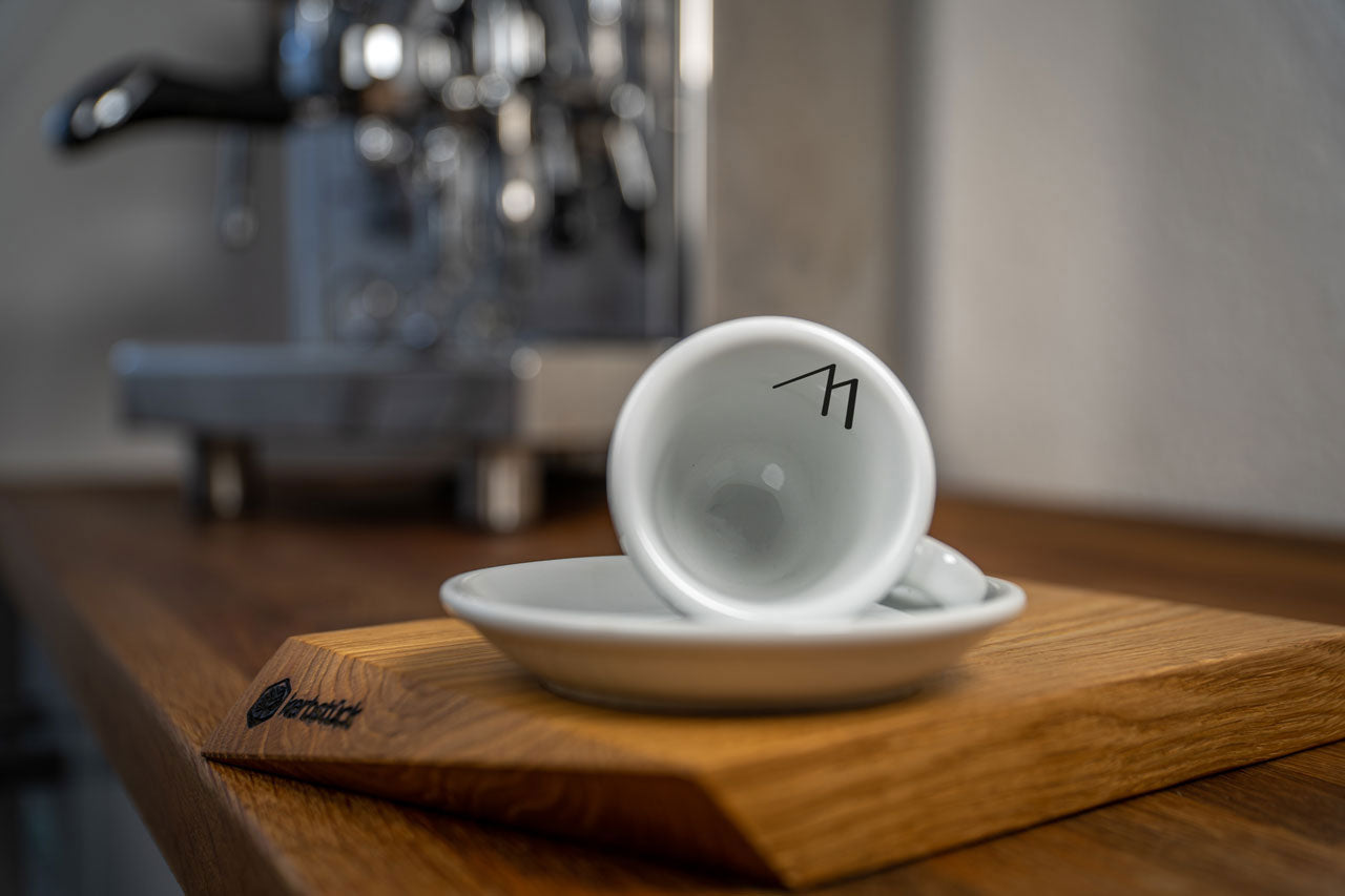 Eine weiße Espresso-Tasse liegt auf der Untertasse. Auf der Innenseite der Tasse sieht man den Druck einer grafischen Darstellung zweier Berggipfel. Im Hintergrund eine glänzende Siebträger-Kaffeemaschine.