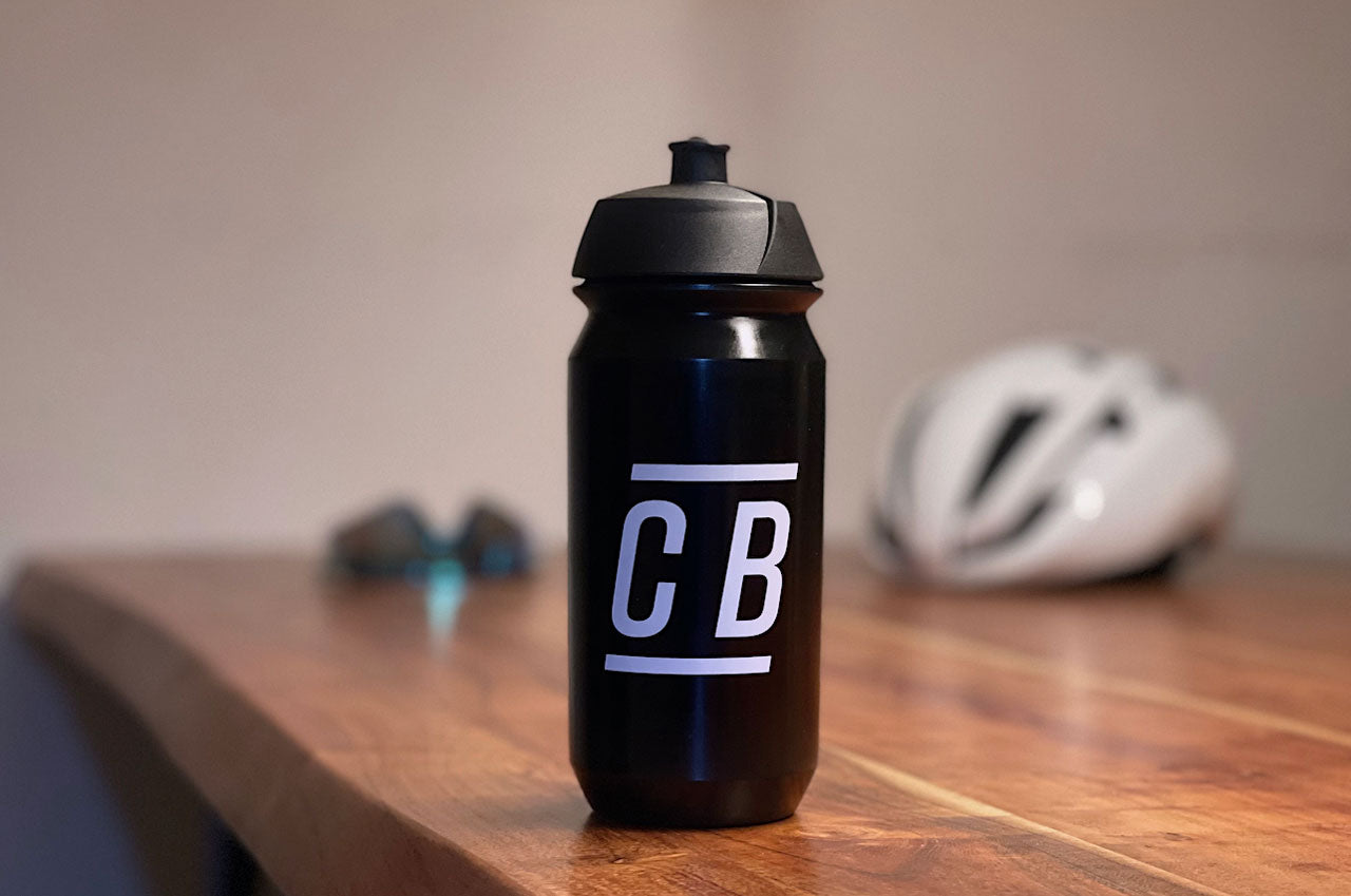 Eine schwarze Fahrrad-Trinkflasche mit dem Logo von CYCLEBEAN auf einem Tisch vor einem verschwommenen Hintergrund.