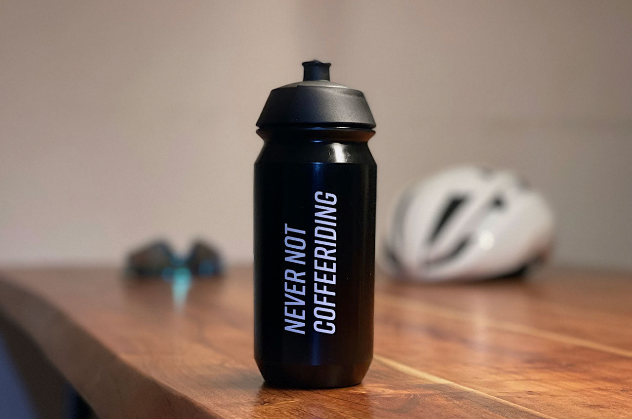 Eine schwarze Fahrrad-Trinkflasche mit dem Spruch "Never not Coffeeriding" auf einem Tisch vor einem verschwommenen Hintergrund.